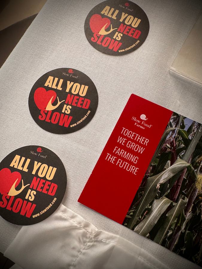 Three round black stickers reading 'All you need is slow' next to a red Slow Food Farms brochure that reads 'Together we grow farming the future' on a white fabric surface